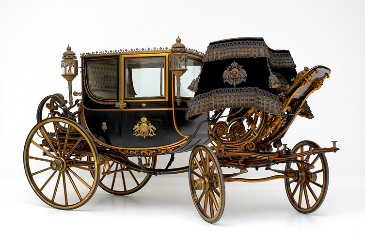 Eine schwarz-goldene verschnörkelte Pferdekutsche mit aufwändigen Details, großen Speichenrädern und dekorativen Laternen vor einem schlichten weißen Hintergrund.