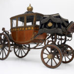 Verschnörkelte antike Pferdekutsche mit goldenen Details, geschlossener Kabine, großen Holzrädern und dekorativem schwarzem Verdeck auf weißem Hintergrund.