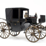 Eine schwarze, alte Pferdekutsche mit vier großen Holzrädern, einem überdachten Fahrgastraum und auf der Vorderseite angebrachten Laternen.