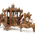 Eine verzierte, goldverzierte königliche Kutsche mit dekorativen Schnitzereien, großen Holzrädern und einer offenen Tür, dargestellt vor einem schlichten weißen Hintergrund.