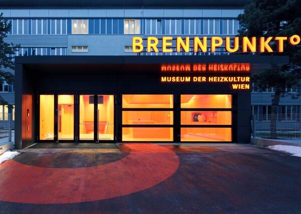Eingang des Brennpunkt° Museum der Heizkultur Wien, mit leuchtend orangefarbener Beleuchtung und Beschilderung über modernen Glastüren.