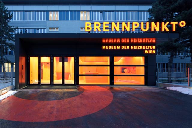Eingang des Brennpunkt° Museum der Heizkultur Wien, mit leuchtend orangefarbener Beleuchtung und Beschilderung über modernen Glastüren.