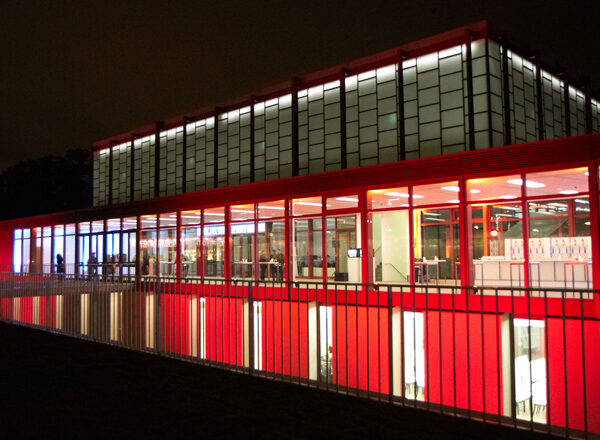 Ein modernes Gebäude mit beleuchteten Glaswänden und roten Akzenten, fotografiert bei Nacht, wobei die Innenbeleuchtung durch große Fenster sichtbar ist.