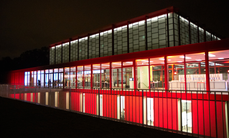 Ein modernes Gebäude mit beleuchteten Glaswänden und roten Akzenten, fotografiert bei Nacht, wobei die Innenbeleuchtung durch große Fenster sichtbar ist.