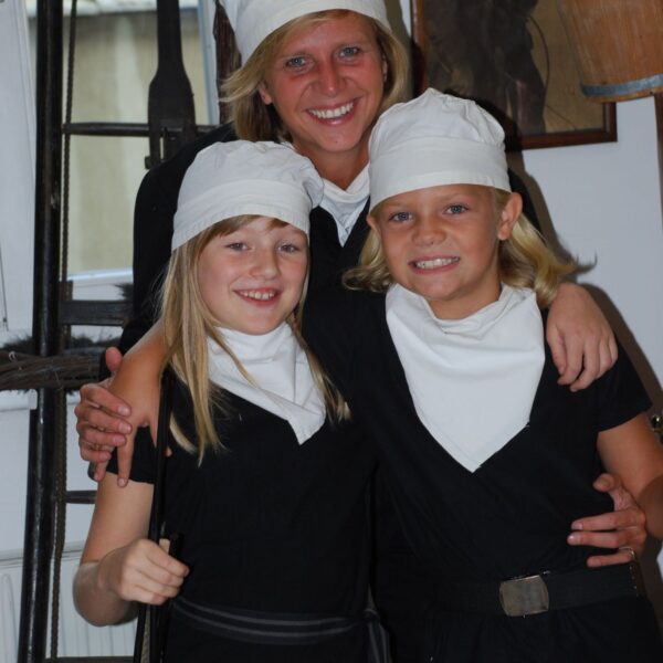 Eine Frau und zwei Kinder, alle schwarz gekleidet mit weißen Hüten und Schals, posieren in einem Haus. Ein Kind hält einen Schornsteinfegerpinsel.