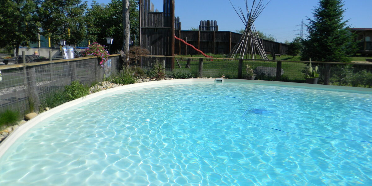 Außenschwimmbad mit klarem Wasser, umgeben von einem niedrigen Zaun, mit einem hölzernen Spielgerät und Tipi-Stangen im grasbewachsenen Hintergrund unter einem klaren blauen Himmel.
