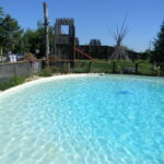 Außenschwimmbad mit klarem Wasser, umgeben von einem niedrigen Zaun, mit einem hölzernen Spielgerät und Tipi-Stangen im grasbewachsenen Hintergrund unter einem klaren blauen Himmel.
