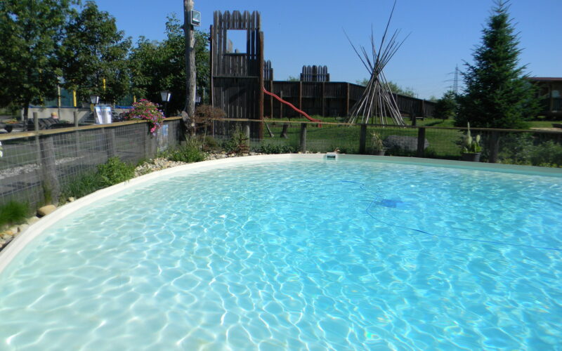 Außenschwimmbad mit klarem Wasser, umgeben von einem niedrigen Zaun, mit einem hölzernen Spielgerät und Tipi-Stangen im grasbewachsenen Hintergrund unter einem klaren blauen Himmel.