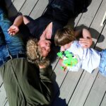 Drei Personen liegen auf einem Holzdeck; eine erwachsene Frau und ein Kind stehen sich gegenüber, wobei das Kind einen grünen Frosch in der Hand hält. Ein erwachsener Mann liegt neben ihnen.