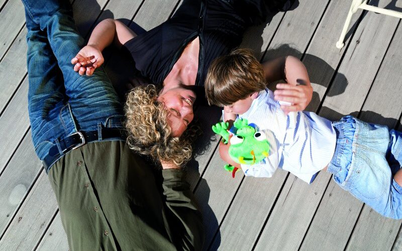 Drei Personen liegen auf einem Holzdeck; eine erwachsene Frau und ein Kind stehen sich gegenüber, wobei das Kind einen grünen Frosch in der Hand hält. Ein erwachsener Mann liegt neben ihnen.