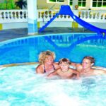 Eine Frau, ein Kind und ein Mann entspannen sich gemeinsam in einem runden Innen-Whirlpool neben einem Pool mit einer blauen Rutsche im Hintergrund.