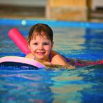Junges Kind schwimmt in einem Pool mit einem Kickboard und Poolnudeln und lächelt in die Kamera.