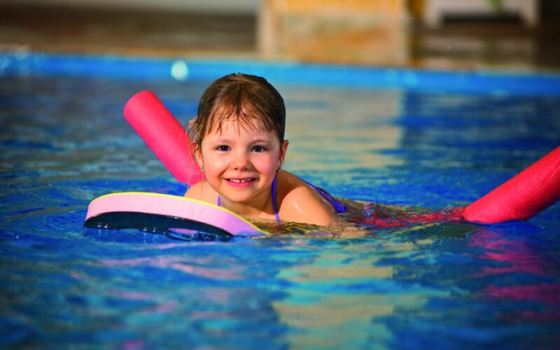 Junges Kind schwimmt in einem Pool mit einem Kickboard und Poolnudeln und lächelt in die Kamera.
