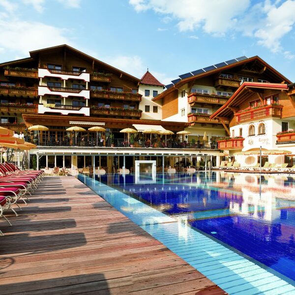 Außenschwimmbad mit roten Liegestühlen, Holzdeck und einem großen Hotelgebäude im alpinen Stil im Hintergrund unter blauem Himmel.