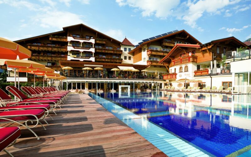 Außenschwimmbad mit roten Liegestühlen, Holzdeck und einem großen Hotelgebäude im alpinen Stil im Hintergrund unter blauem Himmel.