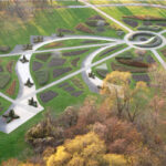 Luftaufnahme eines formalen Gartens mit geometrischen Blumenbeeten, geschwungenen Wegen und einem kreisförmigen zentralen Element, umgeben von Gras und Bäumen.