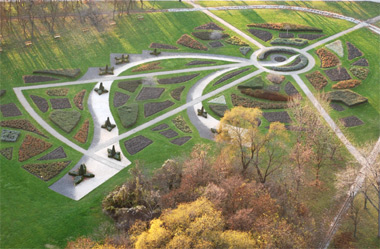 Luftaufnahme eines formalen Gartens mit geometrischen Blumenbeeten, geschwungenen Wegen und einem kreisförmigen zentralen Element, umgeben von Gras und Bäumen.