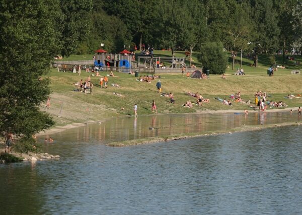 Menschen entspannen und sonnen sich auf einer Wiese am Seeufer in der Nähe eines Spielplatzes, mit Bäumen im Hintergrund und einigen Menschen, die im Wasser waten.