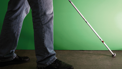 Eine Person in Jeans und schwarzen Schuhen benutzt einen weißen Stock, während sie auf einem Betonboden vor einem grünen Hintergrund geht.