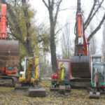 Mehrere Bagger und Baufahrzeuge sind in einer Reihe auf einem unbefestigten Grundstück geparkt, mit Bäumen und heruntergefallenen Blättern im Hintergrund.