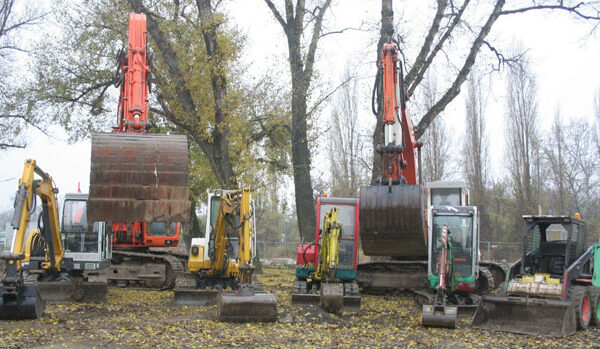Mehrere Bagger und Baufahrzeuge sind in einer Reihe auf einem unbefestigten Grundstück geparkt, mit Bäumen und heruntergefallenen Blättern im Hintergrund.