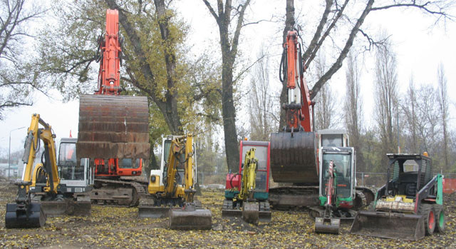 Mehrere Bagger und Baufahrzeuge sind in einer Reihe auf einem unbefestigten Grundstück geparkt, mit Bäumen und heruntergefallenen Blättern im Hintergrund.