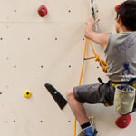 Ein Kind, das einen Klettergurt und einen Helm trägt, klettert an einer Indoor-Kletterwand mit verschiedenen Griffen und Tritten.