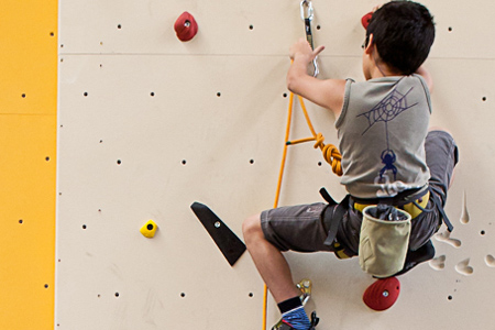 Ein Kind, das einen Klettergurt und einen Helm trägt, klettert an einer Indoor-Kletterwand mit verschiedenen Griffen und Tritten.