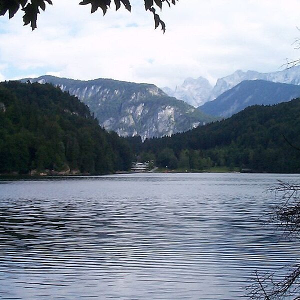 Ein ruhiger See, umgeben von bewaldeten Hügeln und fernen Bergen unter einem teilweise bewölkten Himmel.