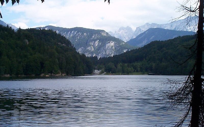 Ein ruhiger See, umgeben von bewaldeten Hügeln und fernen Bergen unter einem teilweise bewölkten Himmel.