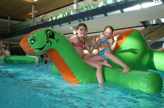Zwei junge Mädchen in Badeanzügen sitzen auf einem großen aufblasbaren grünen Dinosaurier in einem Hallenbad, beide lächeln und zeigen die Daumen nach oben.