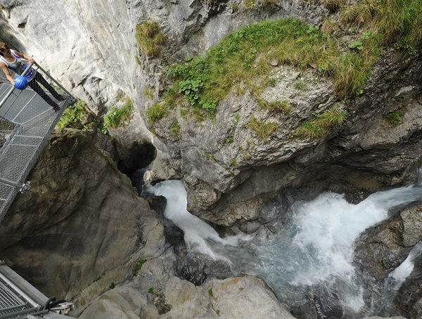 Eine Person steht auf einer Metallbrücke mit Blick auf eine felsige Schlucht mit schnell fließendem, türkisfarbenem Wasser darunter.
