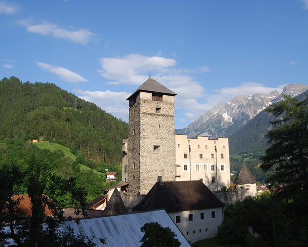 Eine Steinburg mit einem zentralen Turm, umgeben von Bäumen und Hügeln, mit Bergen im Hintergrund unter blauem Himmel.