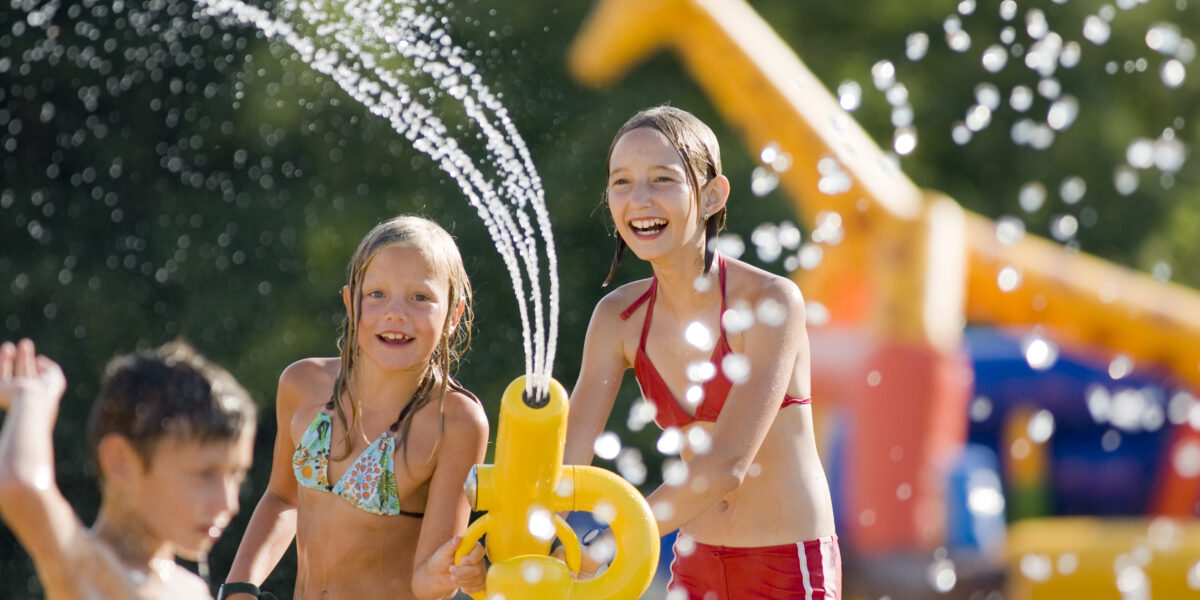 Drei Kinder spielen mit einer Wasserspritzanlage in einem Wasserpark im Freien, wobei im Hintergrund aufblasbare Strukturen und Wassertropfen zu sehen sind.