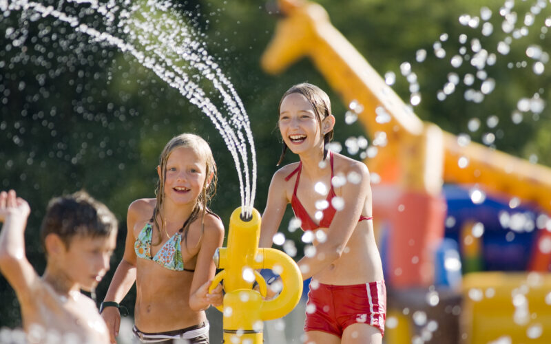 Drei Kinder spielen mit einer Wasserspritzanlage in einem Wasserpark im Freien, wobei im Hintergrund aufblasbare Strukturen und Wassertropfen zu sehen sind.