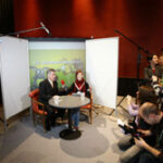 Zwei Personen sitzen während eines Interviews an einem runden Tisch vor einer Kulisse, umgeben von Kameras, Licht und Fotografen in einer Studioumgebung.