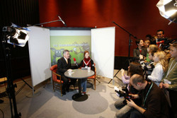 Zwei Personen sitzen während eines Interviews an einem runden Tisch vor einer Kulisse, umgeben von Kameras, Licht und Fotografen in einer Studioumgebung.