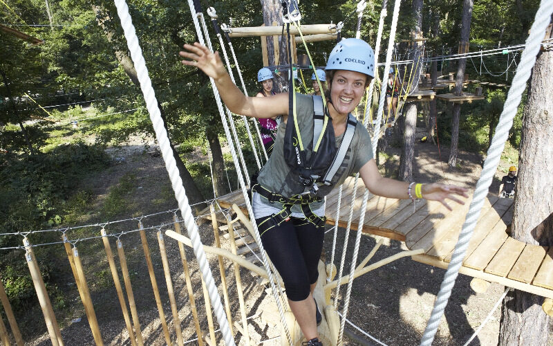 Eine Person, die eine Sicherheitsausrüstung trägt, lächelt und balanciert auf Seilen in einem Outdoor-Abenteuerpark zwischen Bäumen.