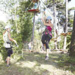 Zwei Frauen mit Helmen und Klettergurten befinden sich in einem Outdoor-Abenteuerpark; eine fährt Seilrutsche, während die andere am Boden steht und zusieht. Bäume und Seilgartenstrukturen sind zu sehen.