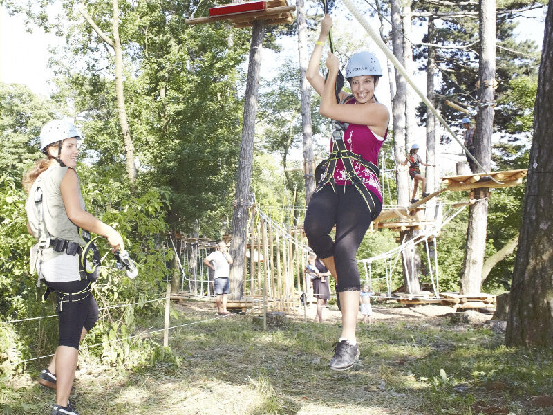 Zwei Frauen mit Helmen und Klettergurten befinden sich in einem Outdoor-Abenteuerpark; eine fährt Seilrutsche, während die andere am Boden steht und zusieht. Bäume und Seilgartenstrukturen sind zu sehen.