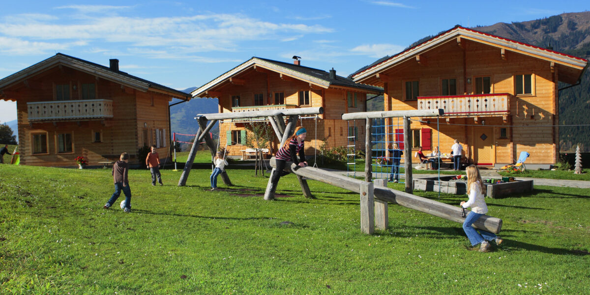 Kinder spielen auf einer Wippe und einem Spielplatz vor drei Holzhäusern im Chalet-Stil auf einer Wiese mit Bergen im Hintergrund.