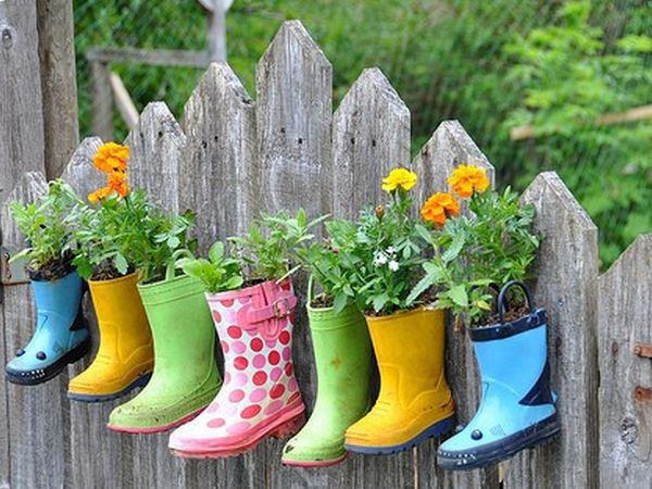 Bunte, mit Blumen gefüllte Gummistiefel hängen an einem Holzzaun und dienen als kreative Pflanzgefäße.