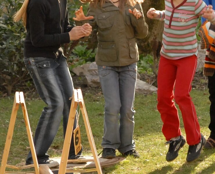 Drei Kinder spielen im Freien, lachen und springen in der Nähe eines hölzernen Hindernisses auf einer Wiese, mit Holzscheiten und gestapeltem Brennholz im Hintergrund.