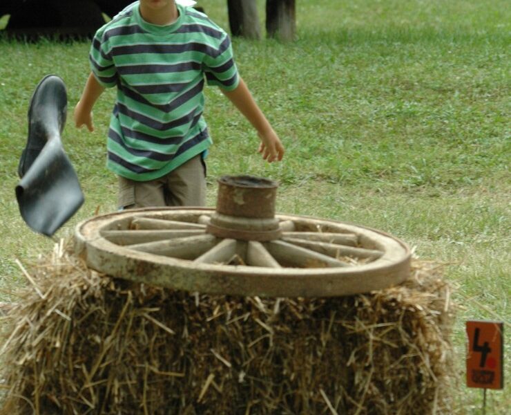 Ein Junge in einem gestreiften Hemd wirft ein Hufeisen auf ein Ziel, das aus einem Wagenrad auf Heuballen in einem grasbewachsenen Außenbereich besteht.