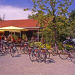 Vor einem Café mit Sitzgelegenheiten im Freien, gelben Sonnenschirmen und Menschen, die an einem sonnigen Tag essen, sind zahlreiche Fahrräder abgestellt.