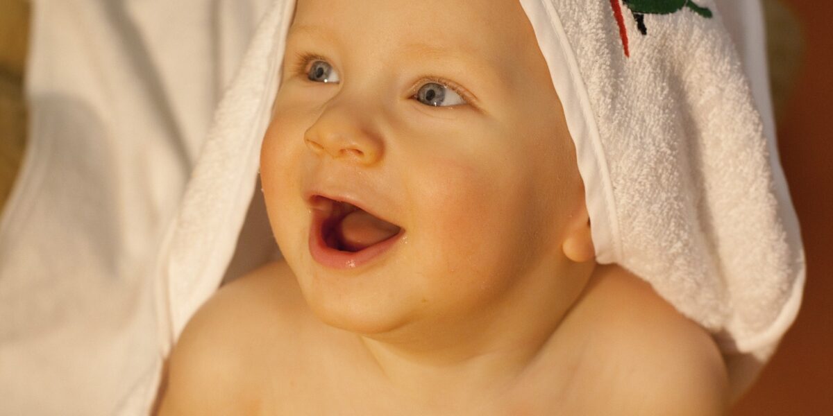 Ein Baby mit einem Handtuch über dem Kopf sitzt lächelnd da und schaut nach oben, im Hintergrund ein Stofftier und eine Decke.
