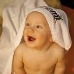 Ein Baby mit einem Handtuch über dem Kopf sitzt lächelnd da und schaut nach oben, im Hintergrund ein Stofftier und eine Decke.