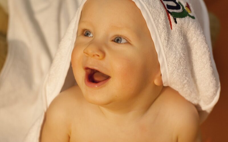 Ein Baby mit einem Handtuch über dem Kopf sitzt lächelnd da und schaut nach oben, im Hintergrund ein Stofftier und eine Decke.