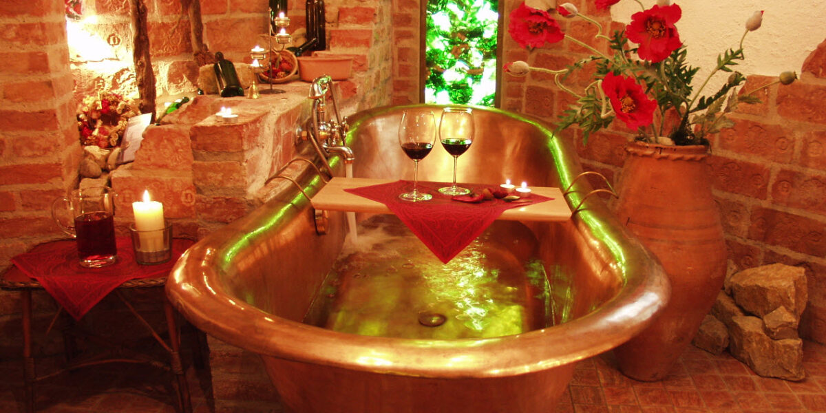 Eine mit Wasser gefüllte Kupferbadewanne, dekoriert mit Kerzen, roten Blumen, Weinflaschen und zwei Rotweingläsern auf einem Holztablett, in einem rustikalen Backsteinzimmer.