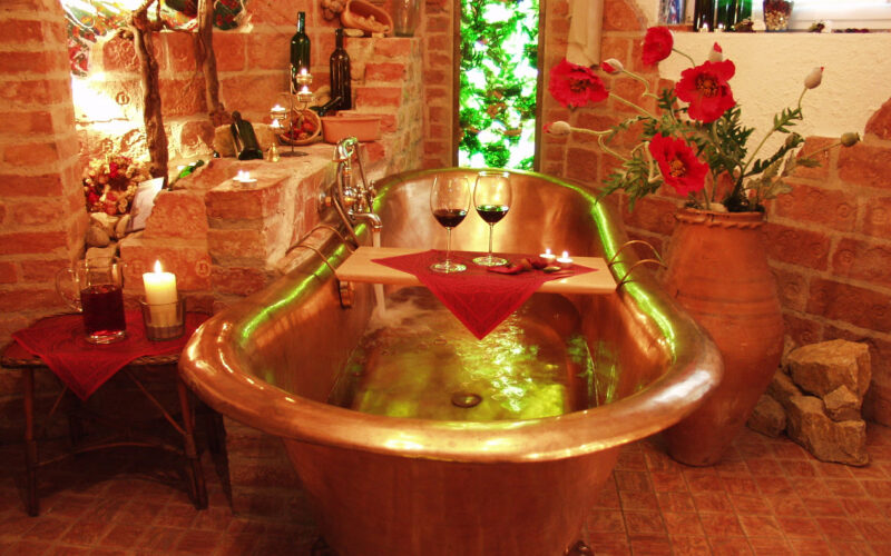 Eine mit Wasser gefüllte Kupferbadewanne, dekoriert mit Kerzen, roten Blumen, Weinflaschen und zwei Rotweingläsern auf einem Holztablett, in einem rustikalen Backsteinzimmer.